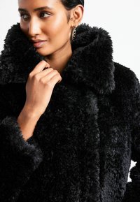 Sort faux fur frakke med høj krave og tekstureret, blød finish. Har brede manchetter og knaplukning. Øreringe er også synlige.