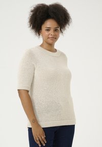 Pull en tricot de couleur beige clair, avec un col rond et des manches courtes à revers. Coupe décontractée avec un ourlet côtelé. Porté avec un jean sombre.