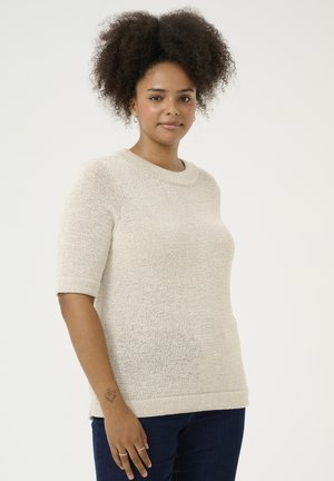 Knit sweater in light beige, featuring a crew neck and short cuffed sleeves. Relaxed fit with ribbed hem. Worn with dark jeans.