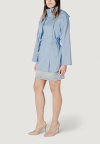 Trench coat impermeabile blu chiaro con maniche lunghe, vita regolabile con cinta, colletto a punta e tasche laterali, abbinato a una gonna di jeans e tacchi beige.