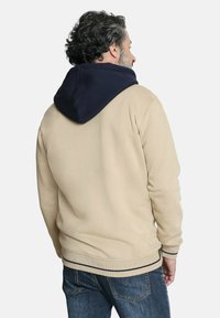 Sudadera beige con una capucha azul marino, que presenta una textura suave, puños acanalados y una franja horizontal a lo largo del dobladillo. Vista trasera mostrada.