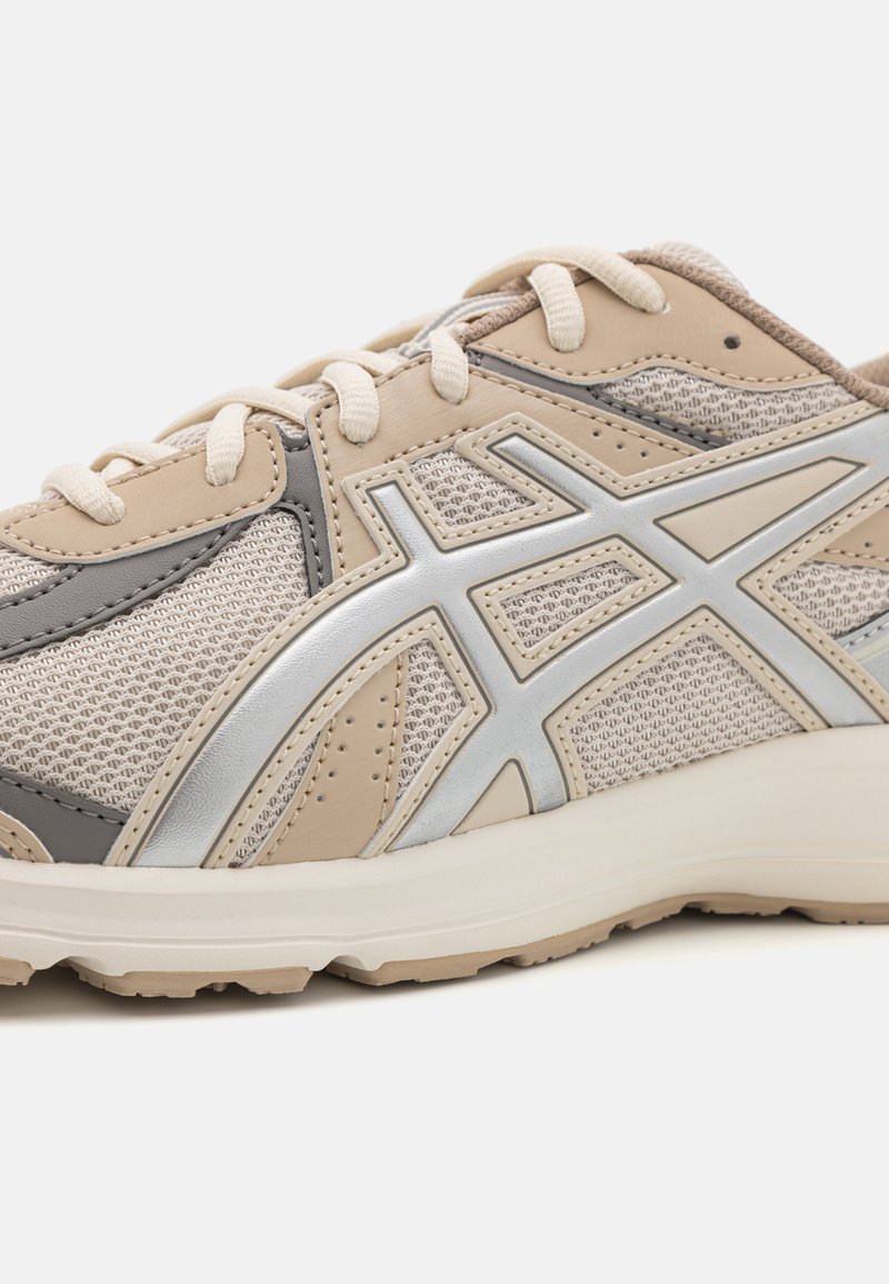 Zapatilla deportiva beige con acentos grises, fabricada con materiales de malla y sintéticos. Cuenta con un logo plateado, una media suela blanca y patrones texturizados en la suela exterior.