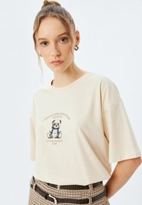 Camiseta de algodón color crema con mangas cortas que presenta un panda de dibujos animados impreso y texto. Combinada con un cinturón a cuadros marrón.