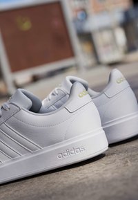Par hvide Adidas sneakers med tre striber og logo på siden og hælen, placeret udendørs på en betonoverflade.