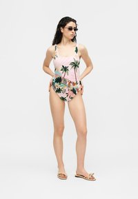 Femme portant un maillot de bain une pièce à imprimé tropical rose, des lunettes de soleil noires et des sandales beige, debout les mains sur les hanches devant un fond uni.