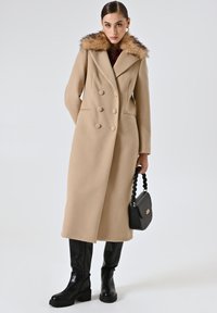 Cappotto beige doppiopetto con collo in pelliccia sintetica, tessuto morbido, silhouette lunga, abbinato a stivali neri al ginocchio e una borsa nera.