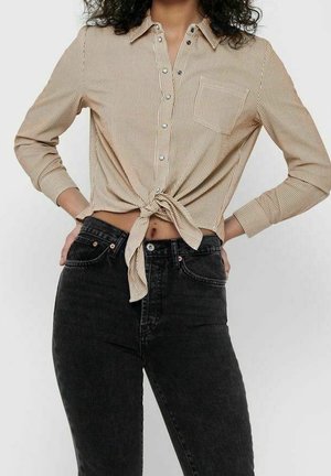 Vrouw draagt een beige gestreept overhemd met knoopjes, gestrikt in de taille, met zwarte high-waist jeans, handen op de heupen.