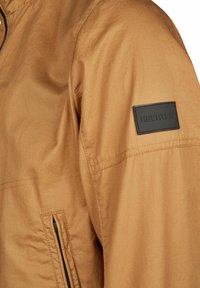 Hellbraune Jacke mit glatter Textur, ausgestattet mit einer Reißverschlusstasche und einem schwarzen Gummipatch mit der Aufschrift "HECHTER" am Ärmel.