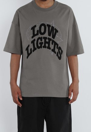Person trägt ein graues, übergroßes T-Shirt mit dem Schriftzug "LOW LIGHTS" und einem dezenten Sternenmuster über einer schwarzen Hose, steht vor weißem Hintergrund.