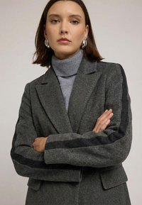 Grå uldfrakke med skræddersyet design, kontrasterende sorte striber på ærmerne og subtile syningsdetaljer. Båret over en grå rullekrave.