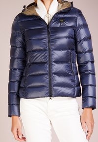 Giacca a vento blu navy con design a trapuntatura orizzontale, chiusura frontale a zip e cappuccio beige. Presenta una patch con logo sulla tasca del petto.