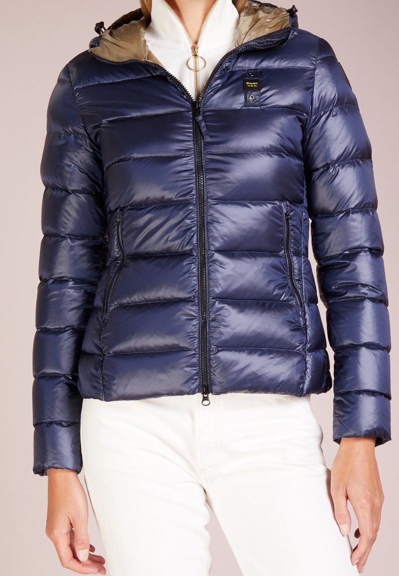 Giacca a vento blu navy con design a trapuntatura orizzontale, chiusura frontale a zip e cappuccio beige. Presenta una patch con logo sulla tasca del petto.