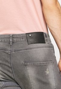 Gray denim jeans featuring a textured finish, a leather logo patch on the back, and a single rear pocket with visible stitching details.