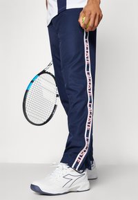 Los joggers azul marino cuentan con rayas laterales con el logo "LACOSTE", combinados con zapatillas deportivas blancas y una raqueta de tenis en la mano. Tejido texturizado.