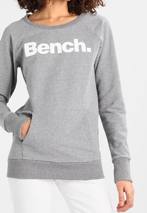 Personne portant un sweat-shirt gris à manches longues avec "Bench." imprimé en blanc sur la poitrine, une main dans la poche et un pantalon blanc.