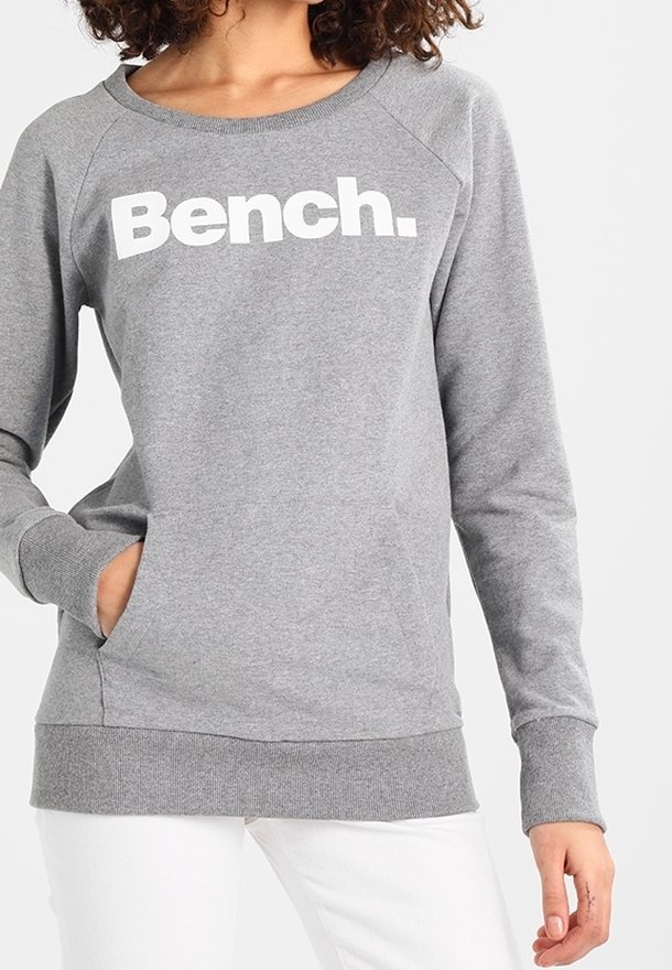 Personne portant un sweat-shirt gris à manches longues avec "Bench." imprimé en blanc sur la poitrine, une main dans la poche et un pantalon blanc.