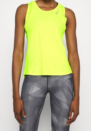 Person trägt ein leuchtend gelbes ärmelloses Sport-Top und graue, gemusterte Sportleggings, steht mit locker hängenden Armen an den Seiten.