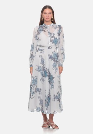 Donna che indossa un abito bianco trasparente a maniche lunghe con stampa floreale blu, chiuso con bottoni sul davanti e sandali bianchi su sfondo semplice.