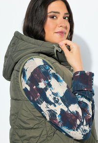 Olivově zelená prošívaná vesta s kapucí, zdobená texturovaným vzorem. Viditelné rukávy košile s modro a vínovo abstraktním potiskem, s detaily na stažení.