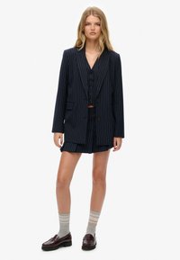 Marineblauwe streeppak met een dubbelgevouwen blazer, bijpassende shorts en een knopenvest. Gedwaald met grijs gestreepte sokken en instappers.
