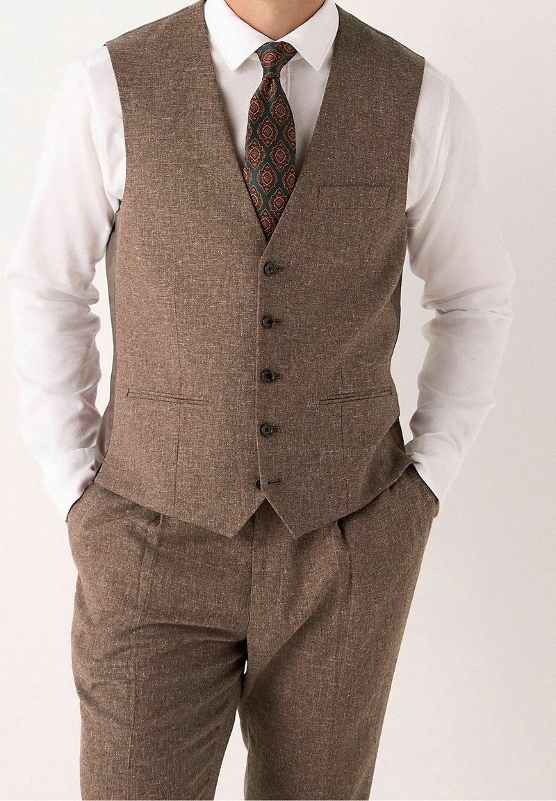 Homme portant un gilet marron boutonné et un pantalon assorti avec une chemise blanche et une cravate verte et marron à motifs, mains dans les poches.
