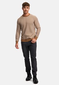 Beige gestrickter Pullover mit strukturiertem Rippengemuster, Rundhalsausschnitt und braunen Akzenten, kombiniert mit dunkelblauen Jeans und schwarzen Schnürstiefeln.