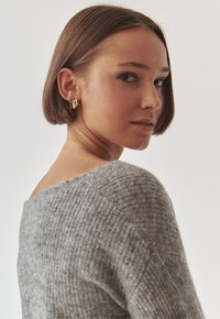 Suéter de punto gris con un escote amplio y un patrón texturizado, combinado con pendientes de aro dorados. La modelo posa con un ligero giro.