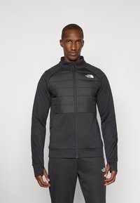 Schwarze Sportjacke mit Reißverschluss auf der Vorderseite, ausgestattet mit einem gepolsterten Brustbereich, gerippten Bündchen und einem kleinen Logo auf der linken Schulter.