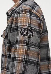 Καρό πουκάμισο φλάναελ σε αποχρώσεις γκρι, μαύρου, λευκού και καφέ με οβάλ ετικέτα "Von Dutch Originals" στον άνω βραχίονα.