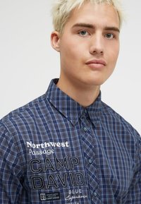 Blau karierte Hemd mit Kragen, verziert mit weißem gesticktem Text und einer Knopfleiste. Aus weichem Stoff mit glatter Textur.