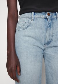 Jeans en denim bleu clair à taille haute, avec un design classique à cinq poches et un ourlet cousu. Comprend une fermeture à bouton marron.