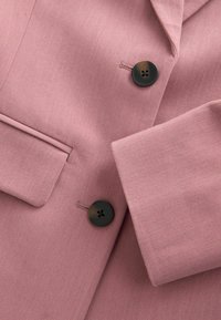 Lyserød blazer med tekstureret stof, hakket revers og to sorte knapper med fire huller på hver, samt en enkelt brystlomme.