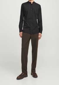 Svart långärmad skjorta med mandarin-krage, knappstängning, i kombination med bruna byxor och mörkbruna skor. Slät tygstruktur.