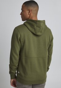 Sweat-shirt à capuche verte avec une texture lisse, présentant un détail de couture à l'arrière et des poignets et ourlet côtelés. Étiquette de marque minimaliste visible.
