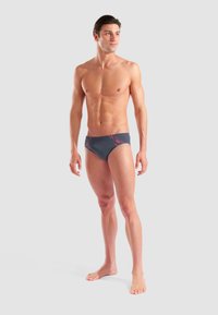 Uomo a torso nudo in costume da bagno grigio con accenti rossi, in piedi a piedi nudi su uno sfondo grigio chiaro uniforme, che guarda a destra.