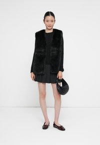 Gilet in pelliccia sintetica nera sopra una maglia nera a maniche lunghe, abbinato a una gonna a pieghe nera aderente, scarpe scure e una piccola borsa nera.
