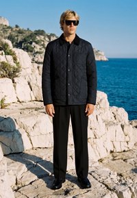 Homme portant une veste matelassée noire, un pantalon noir et des lunettes de soleil, debout sur une côte rocheuse avec l'océan en arrière-plan sous un ciel dégagé.