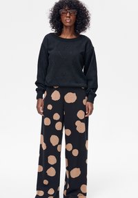 Vrouw met krullend haar en grote bril, gekleed in een zwarte gebreide trui en zwarte broek met wijde pijpen en grote beige polka dots.