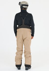 Pantaloni da sci beige con bretelle nere regolabili. Maglietta a maniche lunghe nera sotto. Stivali da neve visibili alla base, design semplice, vestibilità funzionale.