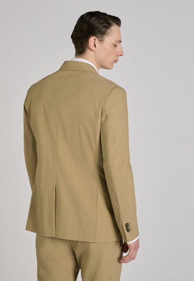 Man met zijn rug naar de camera gekeerd, gekleed in een beige colbert en bijpassende broek, voor een effen lichtgrijze achtergrond.