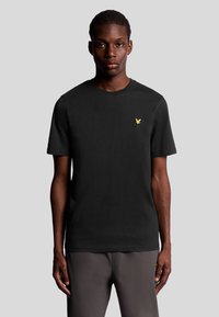 Svart bomullströja med rund hals, korta ärmar och en liten gul örnlogotyp på bröstet. Slät textur, avslappnad passform.