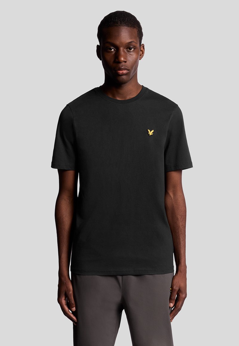 Svart bomullströja med rund hals, korta ärmar och en liten gul örnlogotyp på bröstet. Slät textur, avslappnad passform.