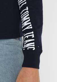 Navy long-sleeve shirt with white "TOMMY JEANS" text on the left sleeve. Features ribbed cuffs and a small logo accent near the hem.