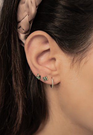 Female ear with three earrings: two green gemstone studs and one small gold hoop with clear stones. Brown hair held back by a beige braided headband.