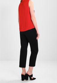 Žena oblečená v bezrukávovém červeném topu, černých zkrácených kalhotách a černých muly s blokovým podpatkem, stojící na jednolitém světlém pozadí.