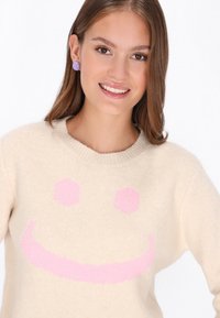 Beiger Strickpullover mit einem rosa Smiley-Gesicht Design. Verfügt über einen runden Halsausschnitt und eine weiche Textur, ergänzt durch runde lilafarbene Ohrringe.