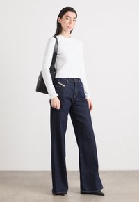 Una donna si trova in un ambiente minimalista, indossando un top bianco a maniche lunghe fitted e jeans a gamba larga scuri, tenendo una borsa nera.