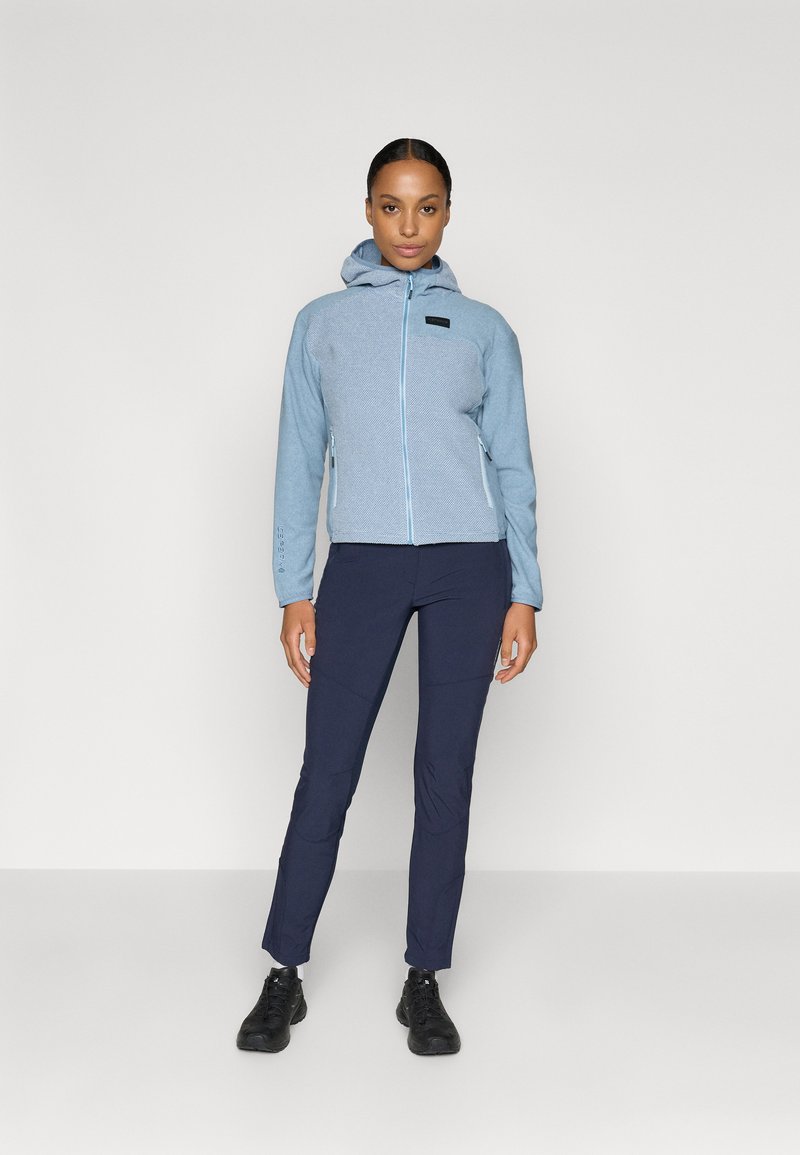 Jasnoniebieska kurtka zapinana na zamek błyskawiczny z kapturem, wykonana z teksturowanej tkaniny, z małym czarnym logo. Zestawiona z ciemnogranatowymi dopasowanymi spodniami i czarnymi butami.