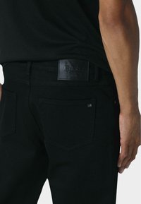 Vaqueros de mezclilla negra con una parche de cuero en la cintura, que presentan un diseño simple, dos bolsillos traseros y sutiles detalles de costura.