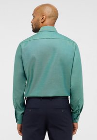 Camicia verde a maniche lunghe con una finitura texturizzata, polsi con bottoni e colletto appuntito, indossata con pantaloni scuri. Vista posteriore.